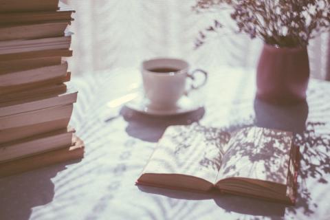 A table with an open book, pile of books, mug and pot of flowers.